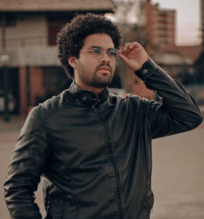 Portrait of a stylish man with afro hair and eyeglasses, wearing a black leather jacket outdoors.