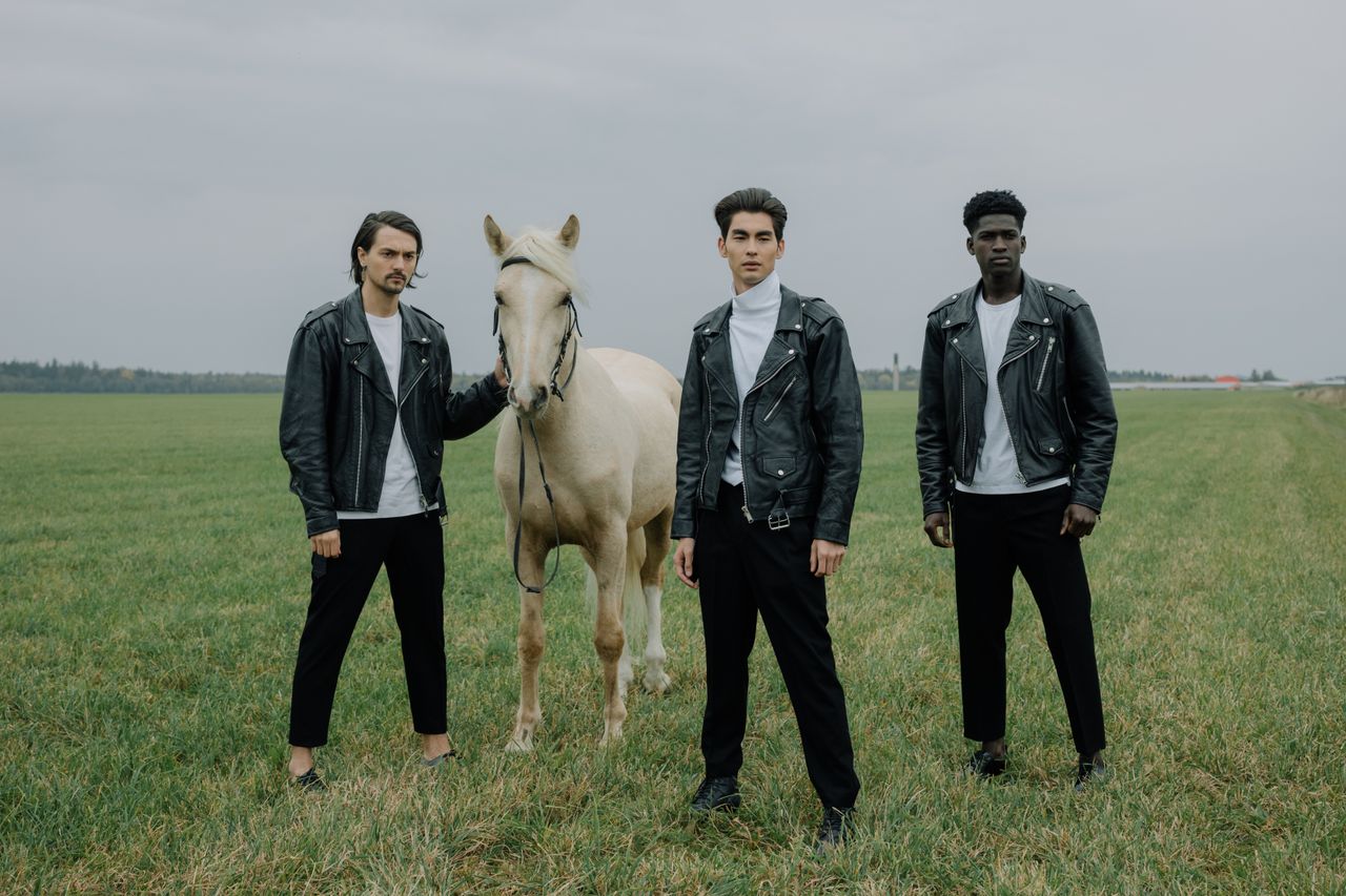 Three men in black leather jackets pose with a horse on a grass field.