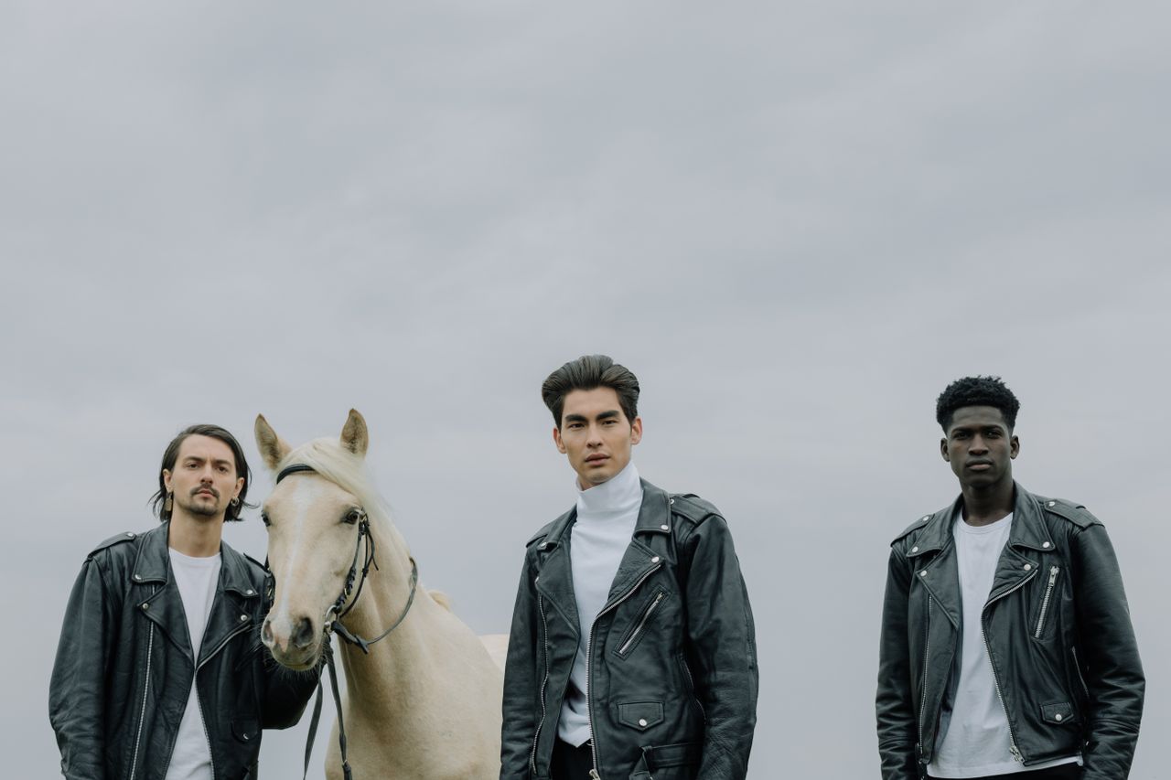 Three men wearing black leather jackets posing with a horse outdoors, showcasing modern fashion.